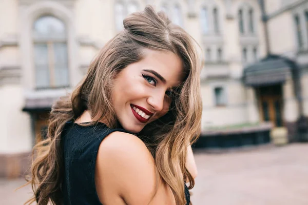 Closeup portrait of pretty girl with long curly hair smiling to camera in city on old building background. She wears black dress, red lips. View from back