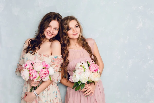 Two beautiful girls stand in a studio and hold bouquets of flowers. They wear light silk dresses. One is blond, and one is brunette. Both have curly hair