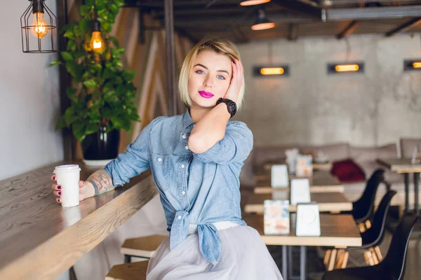 Smiling blond girl sitting in a coffee shop drinking coffee and touching her hair. She wears blue denim shirt and grey tulle skirt. There are wooden chairs and tables in the cafe