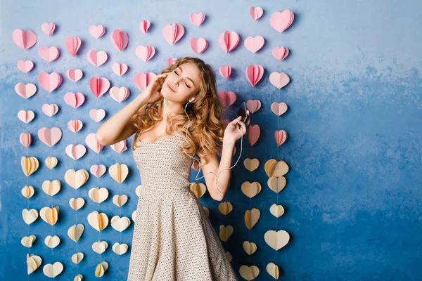 Cute sexy slim girl with blond curly hair standing in a studio with blue background and listenign to music on earphones. She smiles and looks sexy