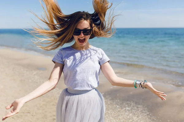 Young pretty girl with long hair is springing on the beach near sea. She has gray T-shirt and skirt. She looks crazy happy