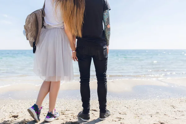 Young couple is standing near sea. He wears black T-shirt and pants. She has long hair, gray T-shirt, skirt and bag. She put head on his shoulder. View from back