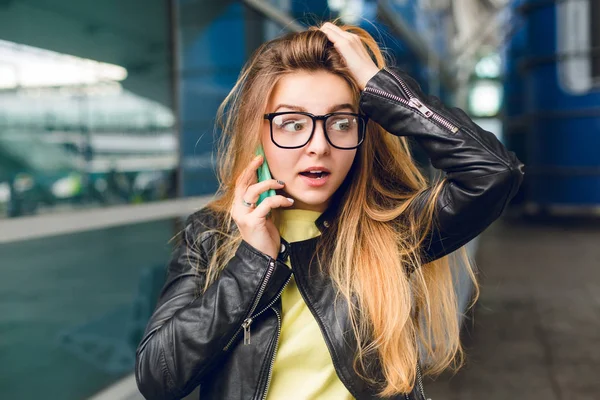Close-up portrait of pretty girl standing outside in airport. She has long hair and black jacket. She is speaking on phone. She looks wonder