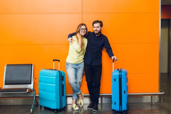 Young couple is standing in airport on orange background between two blue suitcases. Guy with beard in black shirt and pants is hugging girl  with long hair in yellow sweater and jeans