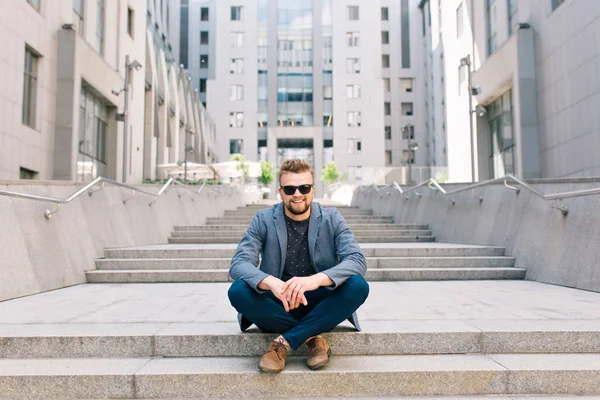 Full-length photo of  man in sunglasses sitting on concrete stairs on  office building background. He wears T-shirt, jacket,  jeans, brown shoes. He looks happy