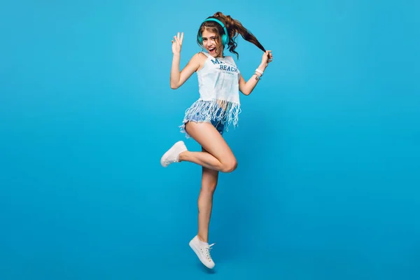 Active girl with long curly hair in tail in jump on blue background in studio. She wears white T-shirt, shorts. She is listening to music with blue headphones