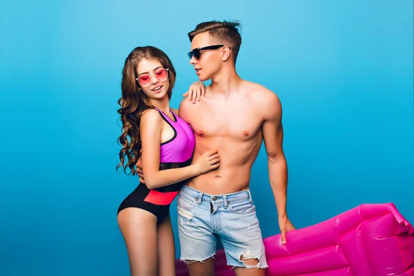 Young couple of hot guy with a bare torso hugging pretty girl with long curly hair in swimsuit on blue  background in studio. They wear sunglasses and he holds pink mattress