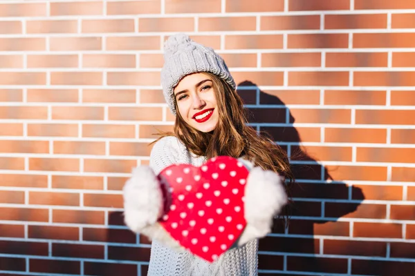 Portrait young girl with long hair in knitted hat, warm sweater on wall background outside. She stretching red heart in gloves to camera.