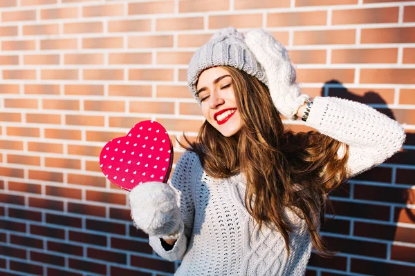 Pretty young girl with long hair in warm sweater and knitted hat on wall background outside. She holds red heart in gloves, looks satisfied with closed eyes.