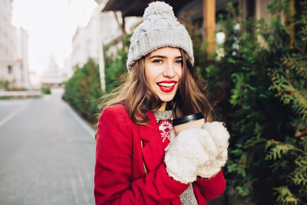 Portrait young girl with long hair in red coat walking on street.  She holds coffee to go in white gloves, smiling to camera.
