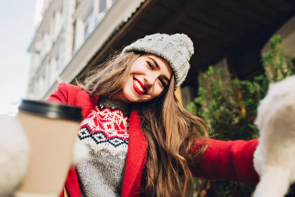 Selfie portrait pretty girl with long hair in red coat on street. She wears knitted hat, white gloves, coffee to go, smiling to camera.