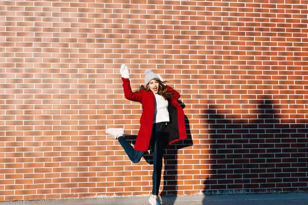 Beautiful brunette girl with long hair having fun on wall background outside. She wears red coat, white gloves and knitted hat.