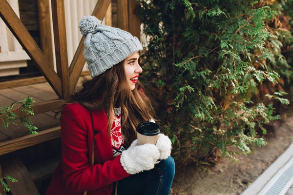 Portrait pretty girl with long hair in red coat sitting on wooden stairs outdoor. She has grey knitted hat, white gloves, holds  coffee and smiling to side.