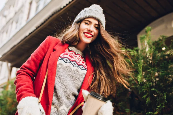 Pretty girl with long hair in knitted hat, red coat walking on street with coffee to go.  She wears white gloves, moving behind camera.