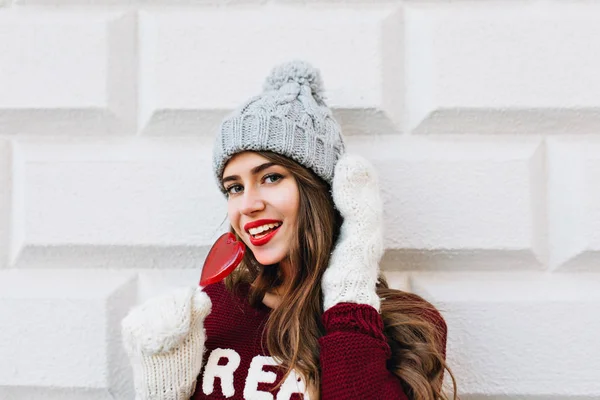 Closeup pretty girl with long hair in marsala sweater on grey wall background. She wears white gloves, grey knitted hat, holds red heart lollipop and smiling to camera.