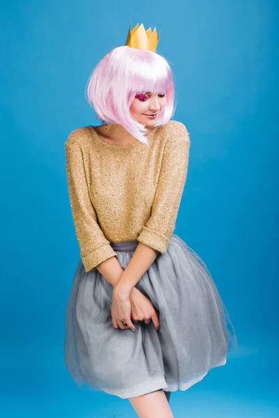 Pretty charming young woman in grey tulle skirt, with pink haircut on blue background. Golden sweater, crown on head, expressing shy emotions, smiling with closed eyes, party, celebration.