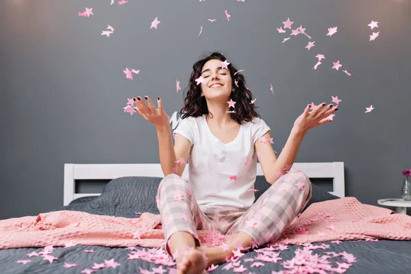 Expressing true positive emotions of young joyful woman in pajamas with cut curly hair having fun in falling pink tinsels on bed in modern apartment. Home cosiness, smiling with closed eyes.