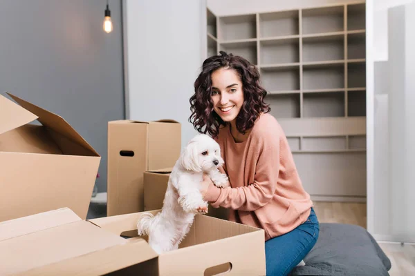 Moving to new modern apartment of joyful young woman finding a little white dog in carton box. Smiling to camera of beautiful model with short curly brunette hair at home comfort.