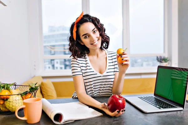 Happy relax time at home of joyful young woman with cut curly hair smiling to camera on table in living room. Laptop with green screen, citrus, apple, magazine, tea, chilling in modern apartment.