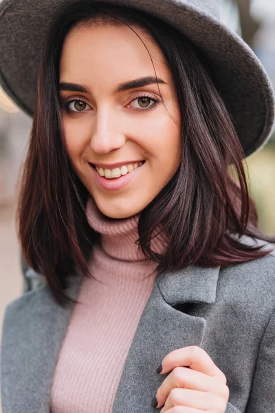 Stylish closeup portrait pretty young  woman with brunette hair walking on street. Grey hat, coat, luxury clothes, elegant outlook, cheerful mood, smiling to camera.