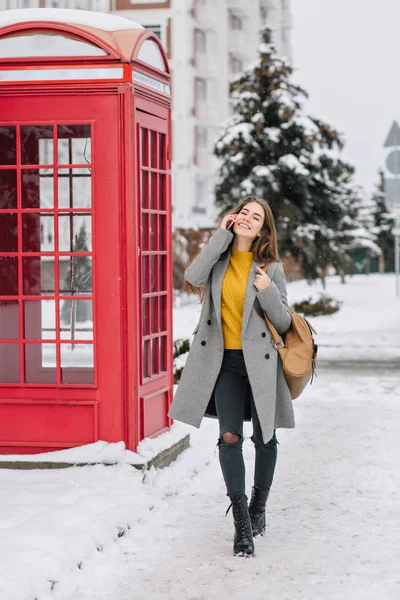 Full-length portrait of cheerful stylish lady talking on phone while walking around winter city. Outdoor photo of pretty caucasian female model with smartphone standing near red call-box..