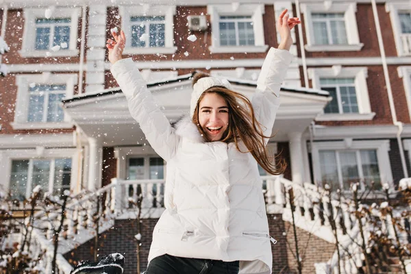 Excited brightful emotions of joyful pretty young woman expressing soround snowfall on street in winter time on house background. Holding hands above, happiness, positivity, joy, winter holidays.