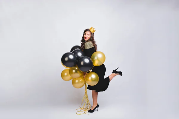 Young amazing woman celebrating new year party on white background. She has black fashion dress, high heels and yellow crown. Brightful carnival, happy birthday, yellow and black balloons, having fun.