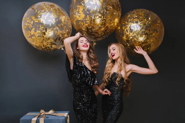 Beautiful ladies dancing with hands up in front of shining helium balloons and smiling. Indoor photo of refined brown-haired birthday girl chilling with friend and laughing..