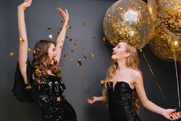Inspired curly pale woman singing with hands up on dark background. Romantic blonde girl in black outfit holding party balloons and looking at friend which dancing under confetti..