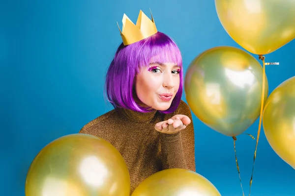 Happy new year party time of attractive young woman sending a kiss to camera surround golden balloons on blue background. Cut purple hair, luxury dress, having fun, birthday celebration.