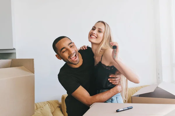 Couple exited about moving in new home, new life. Girl holding keys of new house,  boxes around, man and woman hugging, wearing black top and t-shirt Bright room with white wall on background..