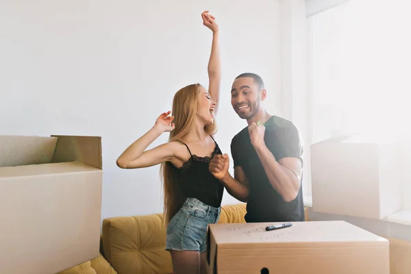 Cheerful man and woman dancing in new apartment, happy about moving, dreams came true, bought new flat, beginning of new life. Girl wearing denim shorts and black top, man - black t-shirt..