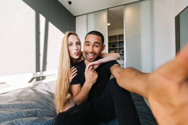 Funny moments of cute couple having fun, making selfie on bed at home in modern appartment. Woman with long blonde hair, true emotions, in love, wife, husband, relationship.