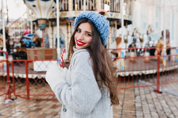 Portrait of blissful long-haired female model in white gloves holding candy cane in amusement park. Pretty girl in blue hat celebrating christmas and posing near carousel..