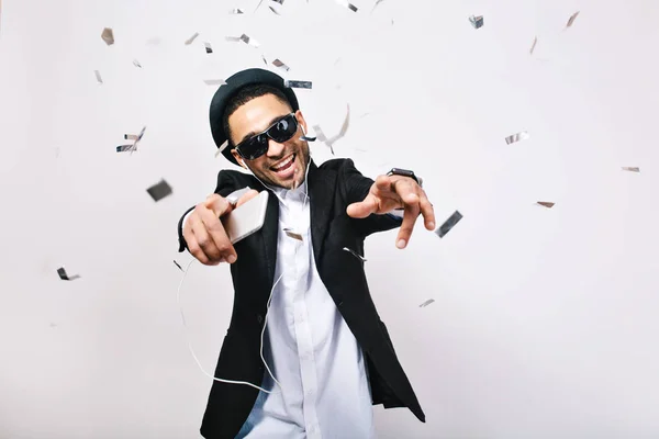 Happy weekends, crazy party time of excited handsome guy in suit, hat, black sunglasses having fun in tinsels on white background. Fashionable look, singing, dancer, happiness, expressions, enjoying.