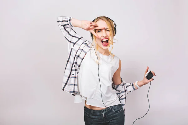 Positive portrait of joyful energy girl with blonde hair in casual outfit listening to music with big headphones. She is dancing and holding smartphone. Isolated. .
