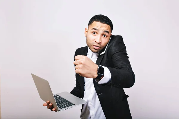 Portrait astonished handsome young man in white shirt, black jacket holding laptop, speaking on phone on white background. Office worker, modern job, true emotions.