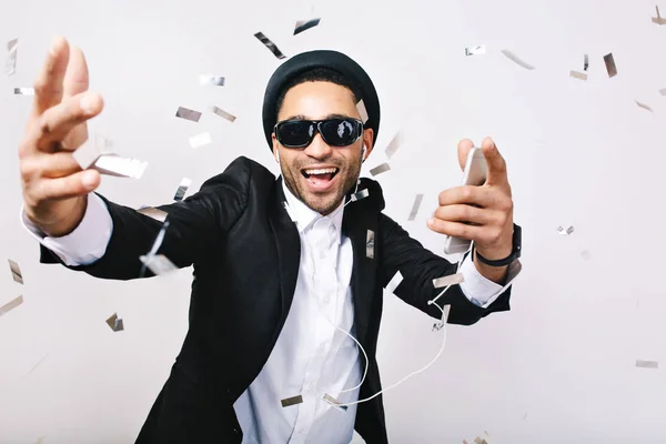 Happy excited party time of joyful handsome guy in hat, suit, black sunglasses having fun in tinsels on white background. Listening to music through headphones, celebrating, singer, super star.