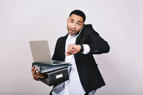 Busy hard-working guy in white shirt and black jacket talking on phone, holding folders, laptop, looks astonished at watch on white background. Office worker, modern technology, career.