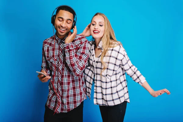 Joyful handsome man listening to music in headphones with closed eyes on blue background. Pretty young woman with long blonde hair having fun with young guy enjoying music. Cheerful mood, smiling.