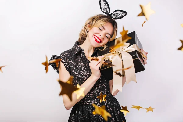 Crazy party time of beautiful women in elegant black dress with gift box celebrating birthday,sparkling gold confetti, having fun, dancing. Emotion face,red lips, eyes closed