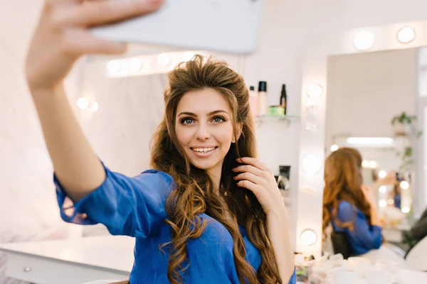Attractive young joyful woman in blue shirt with long brunette hair expressing positive emotions, making selfie in beauty salon. Gorgeous model, preparing to party, satisfaction, smiling.