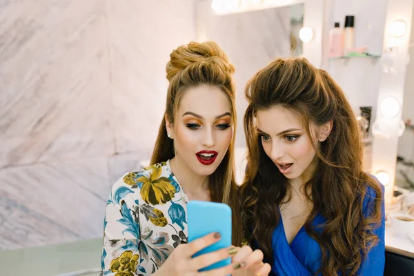 Portrait two astonished pretty women looking at phone in beauty salon. Having fun, gossip girls, preparing to party,  friends together, true emotions, professional makeup, coiffure, stylish models.