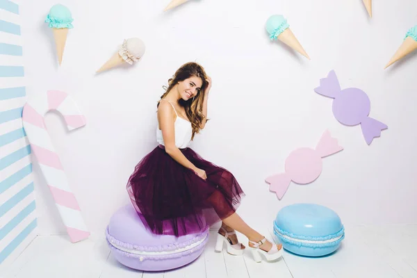 Sweet happy summer time of attractive fashionable young woman in tulle skirt sitting on huge macaron on white background. Pastel colors, sweets, delicious, enjoying, happiness, smiling, relaxing.