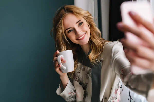 Pretty young woman with wavy blonde hair taking selfie sitting next to the window with cup of morning coffee, tea. She wearing silk pajama. Turquoise wall on background. .