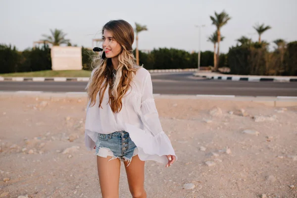 Slim long-haired girl in vintage white blouse walking on the sand with exotic palm trees on background. Charming young woman with cute hairstyle spending time outside and looking away with interest.