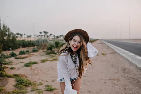 Laughing girl in trendy brown hat and white blouse having fun standing on sand next to highway. Portrait of charming long-haired young woman dancing outside and posing with smile, enjoys vacation.