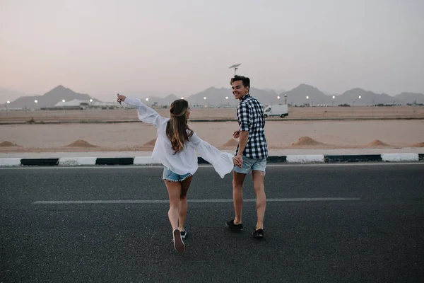Guy in denim shorts and long-haired girl in trendy blouse running across the road and enjoys mountain view. Laughing young couple holding hands walking on highway and having fun outside in summer .