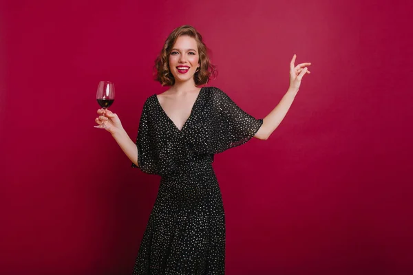 Amazing woman in vintage dress dancing and waving hands on bright background. Smiling european female model with elegant hairstyle holding glass of tasty wine..