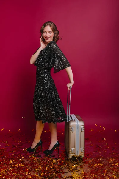 Slim woman in high heel shoes packed her clothes and waiting for travel. Full-length portrait of stylish caucasian female model holding suitcase, standing on confetti..